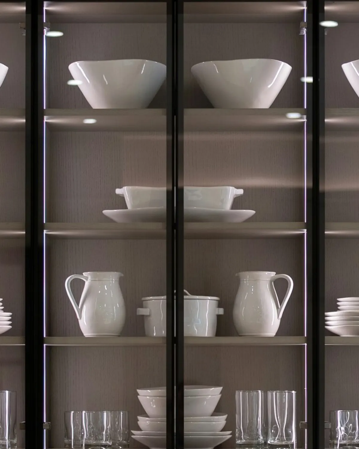 Wandkast met zwarte stalen kaders en helder glas, met wit servies op verlichte planken
